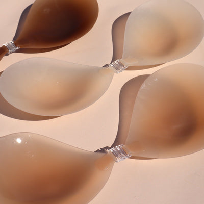 Product shot of reusable and sticky Dark tint, Light tint, and Medium tint Silicone Stick-on Bras laying flat against light pink background, close-up shot showing the clasps and thin edges on each bra